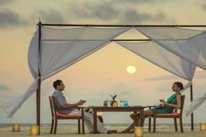 Ein Paar sitzt unter einem Baldachin am Strand und genießt ein romantisches Abendessen bei Sonnenuntergang im Angsana Velavaru.