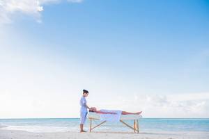 Eine Massage an einem Strand, durchgeführt von einem Therapeuten, mit einem Klienten, der auf einer Massageliege liegt, im Kandima Maldives Hotel.