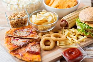 Eine Auswahl an ungesundem Essen wie Pommes, Pizza, Burger, Chips und Soda auf einem Holztisch.