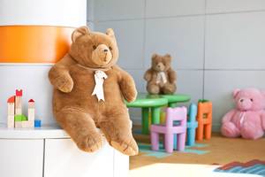 Ein großer brauner Teddybär sitzt auf einem weißen Schrank im Kinderzimmer neben bunten Spielzeugen.