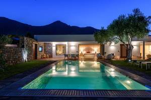 Außenbereich des Elounda Beach Hotel & Villas bei Nacht, mit beleuchtetem Swimmingpool und Blick auf Berge.
