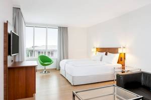 Innenansicht eines Hotelzimmers im NH Hamburg Altona mit zwei Betten, Fernseher, Schreibtisch und grünem Stuhl.