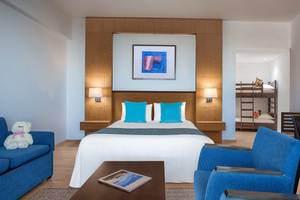 Innenansicht eines Zimmers im Mediterranean Beach Hotel in Zypern mit einem Bett, zwei blauen Kissen, einem Sofa, einem Tisch, einem Teddybär und einem Bild an der Wand.