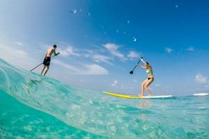 Zwei Personen surfen im Meer bei klarem Himmel mit kleinen Wellen und einem Boot im Hintergrund.