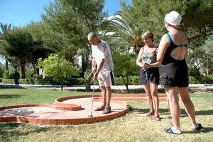 Drei Personen spielen Golf auf einem Minigolfplatz im Freien mit Bäumen und Gebäuden im Hintergrund.