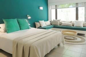 Ein türkisfarbenes Zimmer mit einem großen Bett, grünen Kissen und weißen Decken, einem weißen Stuhl und einer Couch in Blau. Neben dem Bett befindet sich ein Tisch mit einer Tischlampe.