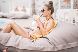 Frau im roten Bikini sitzt mit einem Getränk auf einem grauen Loungesofa am Pool der Ibiza Sun Apartments.