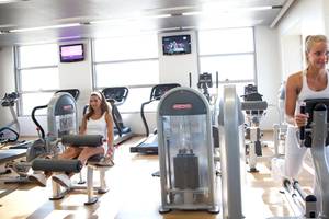 Zwei Frauen trainieren in einem Fitnessstudio, eine auf einem Stepper, die andere auf einem Beinpressegerät.