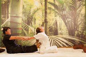 Zwei Frauen praktizieren Yoga in einem Hotel-Spa-Raum mit grüner Wand und tropischem Fototapete.