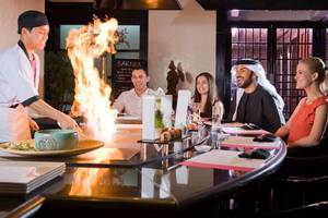 Gäste sitzen an einem Tisch im Millennium Plaza Downtown Sushi Bar und beobachten den Koch, der ein Gericht mit Feuer zubereitet.