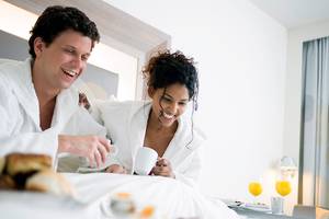 Ein Paar genießt Frühstück im Bett im Novotel London Blackfriars, beide tragen weiße Bademäntel, mit Kaffee, Orangensaft und Frühstücksbuffet im Hintergrund.