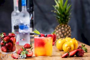 Ein Glas mit fruchtigem Cocktail, dekoriert mit Beeren und Zitrone, vor einem Hintergrund von weiteren Früchten und Vodka-Flaschen.