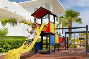 Kinder spielen auf einem bunten Spielplatz im Freien, im Hintergrund steht das H10 Estepona Palace Hotel.