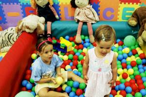 Zwei Mädchen spielen in einem Ballbad mit bunten Bällen, während Puppen und Spielzeuge im Hintergrund stehen.