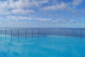 Ein blauer Pool mit Glasbarrieren bietet Aussicht auf das Meer und den Himmel mit Wolken.