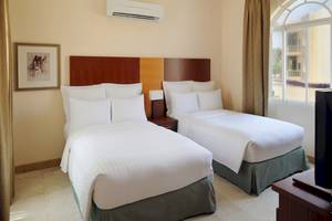 Zwei geräumige Betten in einem Hotelzimmer mit weißem Bettzeug und Kissen, neben einem Nachttisch mit Lampe, Fenster und Klimaanlage.