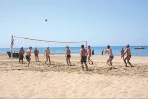 Gruppe von Menschen spielt Volleyball am Strand, im Hintergrund ist das Meer zu sehen.
