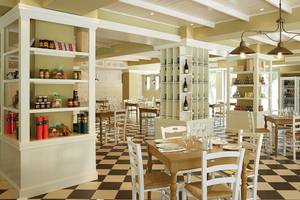 Innenansicht des MarBella Corfu Restaurants mit schachbrettartigem Boden, weißen Möbeln und Regalen mit Produkten.
