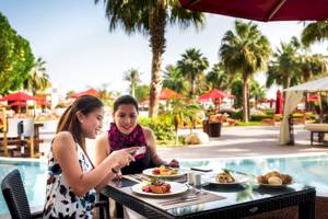Zwei Frauen am Pool sitzend und Essen genießend im Khalidiya Palace Rayhaan by Rotana