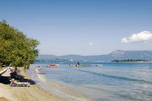 Strandszene am Louis Kerkyra Blue Hotel mit Liegestühlen, Bäumen und Bergen im Hintergrund.