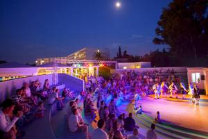 Menschen sitzen auf Stufen und beobachten eine Aufführung auf einer Bühne, Louis Kerkyra Blue Hotel bei Nacht.