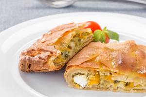 Zwei Stücke Spinat und Feta in Filoteig auf einem weißen Teller mit einer Tomate und Basilikum.