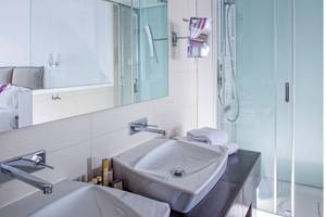 Badezimmer mit einem großen Spiegel und zwei Waschbecken, reflektiert ein Bett und eine Dusche.