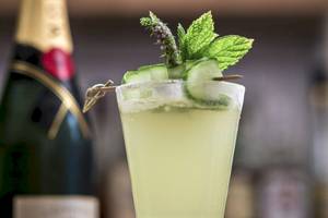 Ein Glas mit einem Cocktail, garniert mit Gurkenscheiben und Minze, vor einer Flasche Champagner.