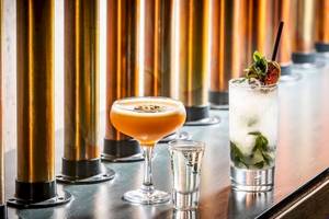 Drei Cocktails auf einer Bar mit goldenen Säulen, darunter ein Orangen-Cocktail, ein Mojito und ein Shot.