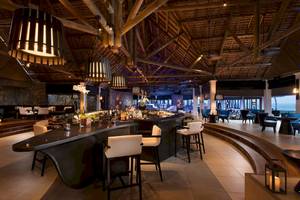 Innenansicht der Bar im Constance Ephelia Mahe Seychelles mit hohen Holzdecken und weißen Stühlen.