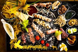 Frühstücksbuffet mit Pasta, Fisch, Schinken und Gemüse auf einem schwarzen Teller.