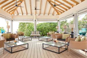 Lounge-Bereich des Amari Havodda Maldives mit Holzboden, modernen Möbeln und großen Fenstern, die einen Blick auf die Außenlandschaft gewähren.