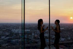 Zwei Frauen in Abendkleidern trinken Wein und genießen den Panoramablick auf Bangkok vom AVANI+ Riverside Bangkok.