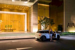 Eine Frau steht neben einem weißen Mini Cooper vor dem AVANI+ Riverside Bangkok Hotel.