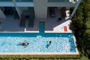 Luftaufnahme von zwei Personen, die im Pool schwimmen, vor einem Hotel mit Terrasse und Liegestühlen.