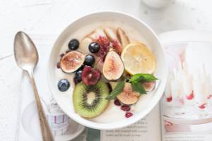 Ein weißer Schüssel mit Joghurt, gefüllt mit verschiedenen Früchten wie Feigen, Blaubeeren, Kiwis und Pomegranate, dazu ein Löffel und ein Buch.