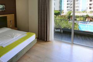 Dyrrah Hotel Zimmer mit Balkon und Poolblick zeigt ein gemütliches Bett, zwei weiße Kissen, eine grüne Decke, eine Glastür und einen Swimmingpool.