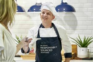 Koch mit Apron, das 'Wir servieren verantwortungsvoll beschafften Fisch' trägt, gibt einer Frau Essen im Iberostar Cristina Hotel.