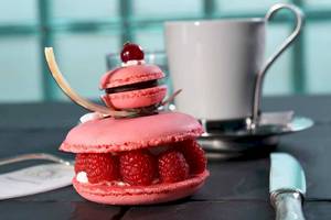 Ein rosa Marmorkuchen mit Himbeeren und einer Kirsche auf einem Tisch neben einer Tasse und einem Messer.