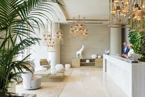 Empfangsbereich des Ikos Dassia Hotels mit modernem Design, weißen Möbeln, Pflanzen und einer Statue.
