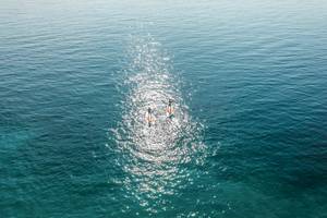Zwei Personen auf Surfbrettern im kristallklaren Wasser vor einer blauen Himmelslandschaft.