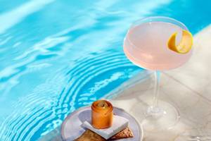 Ein Rosa Cocktail in einem Glas mit einer Zitronenscheibe steht am Rand eines Pools neben einem Teller mit einem gewickelten Keks und einem Marshmallow.