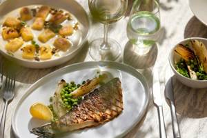 Ein Teller mit gegrilltem Fisch, Erbsen und Sellerie auf einem Tisch mit weiteren Gerichten und Getränken.