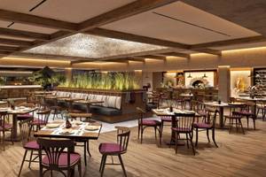 Innenansicht des Restaurants im Ikos Porto Petro Hotel mit Holztischen und Stühlen, Pflanzen und hoher Decke.