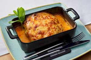 Lasagne im schwarzen Schälchen auf einem blauen Teller serviert, mit Messer, Gabel und Minzeblatt.