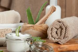 Ein Glas mit Aloe-Vera-Creme, umgeben von einem Holzmörser, einem Schwamm, einer Aloe-Vera-Pflanze und einem gerollten Handtuch.