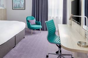 Ein elegantes Hotelzimmer im Leonardo Hotel London Croydon mit einem großen Bett, zwei grünen Stühlen und einem Schreibtisch.