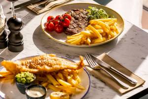 Zwei Teller mit Steak und Pommes sowie Fisch und Pommes auf einem Marmortisch im Leonardo Hotel London Croydon.