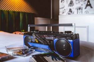 Ein Boombox auf einem Bett im ReMIX Hotel neben Musikkassetten und einem Kissen.