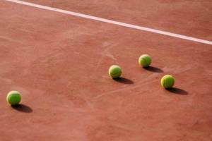 Vier Tennisbälle auf einem roten Tennisplatz neben einer weißen Linie.
